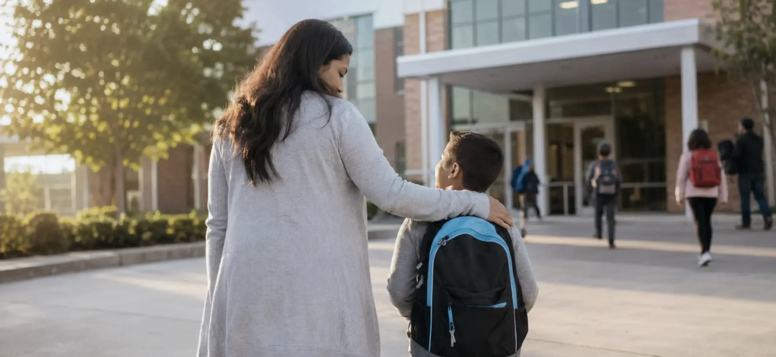 A parent walks their child into school on the first day, holding hands and smiling reassuringly.